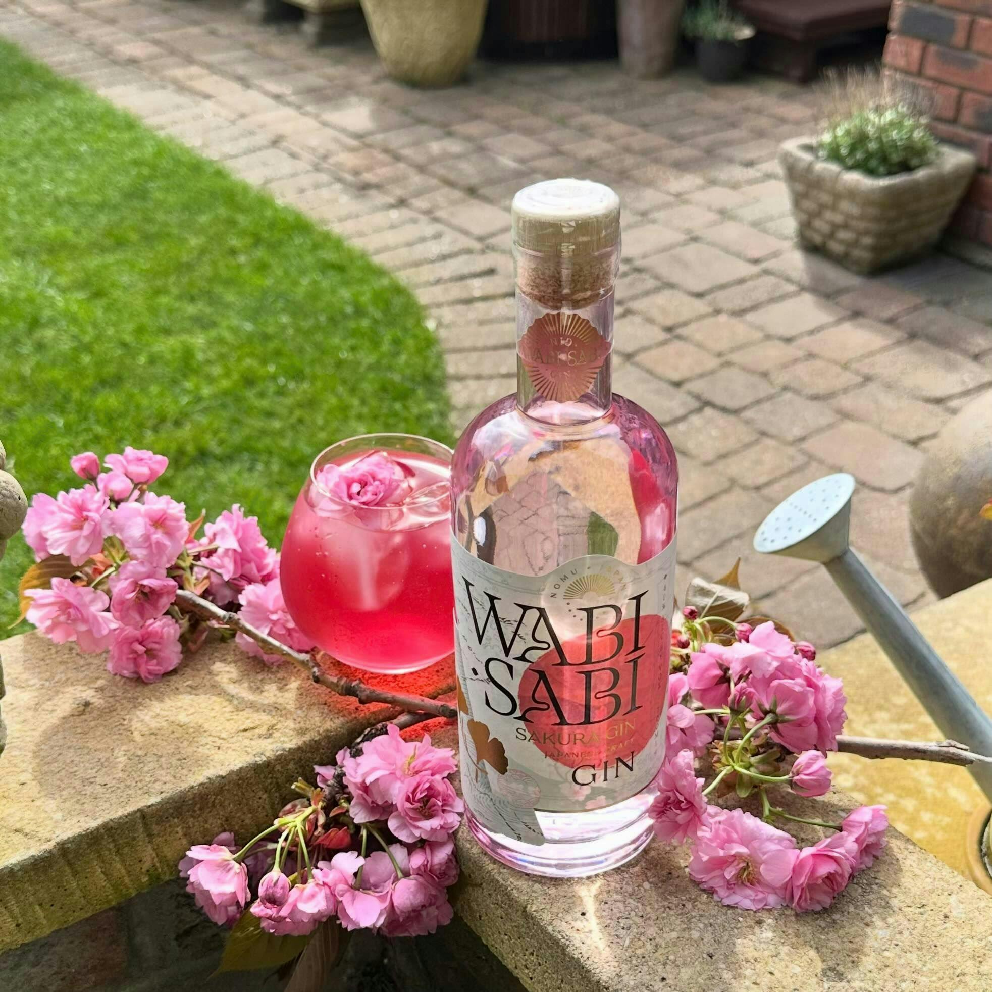 A white and pink labelled bottle of Wabi Sabi gin with a pink cocktail in the garden in the background and some cherry blossom next to the bottle and cocktail