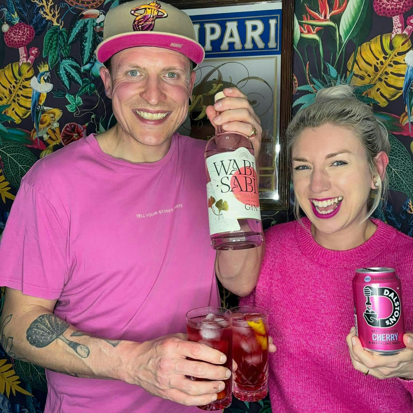 Two friends in pink t-shirts and jumpers with a pink cocktail each and a bottle of Wabi Sabi gin and cherry soda looking happy