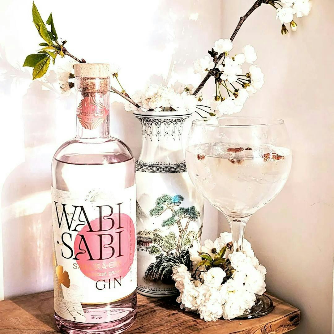 A white and pink bottle of gin from Wabi Sabi with a ceramic vase and cherry blossom in the background and a clear gin glass with a gin and tonic in.